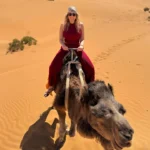 Frau in rotem Gewand reitet auf einem Kamel durch die Wüste bei tief stehender Sonne