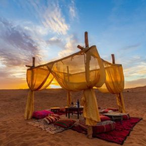 Stimmungsvoll eingerichtetes Wüstencamp mit Baldachin, Teppichen und Sitzkissen bei Sonnenuntergang