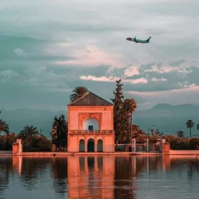 Menara-Garten in Marrakesch mit Pavillon und reflektierendem Wasserbecken, darüber ein fliegendes Flugzeug bei Sonnenuntergang