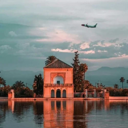 Menara-Garten in Marrakesch mit Pavillon und reflektierendem Wasserbecken, darüber ein fliegendes Flugzeug bei Sonnenuntergang