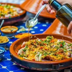 Traditionelle marokkanische Tajine-Gerichte mit Brot und Oliven auf einem blauen Tisch, darüber wird Öl gegossen