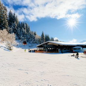 Verschneite Berghütte mit Skifahrern auf einer Piste umgeben von schneebedeckten Bäumen und Alpenlandschaft