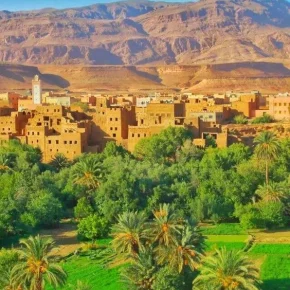 Traditionelle Lehmbauten einer Oasenstadt im Süden Marokkos mit grünen Palmen und landwirtschaftlichen Feldern vor einer Bergkulisse