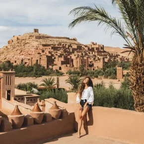 Frau posiert vor der Lehmsiedlung Aït-Ben-Haddou in Marokko mit Palmen im Vordergrund und klarer Sicht auf die Kasbah