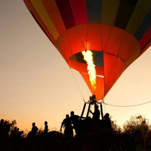 Heißluftballon beim Start bei Sonnenaufgang mit Menschen im Vordergrund und Flamme im Balloninneren